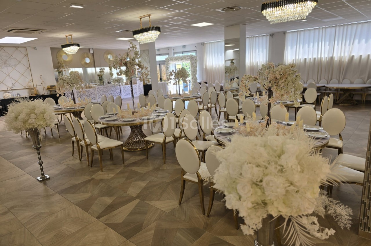 Salle de réception élégamment décorée avec des tables rondes, des chaises blanches et des compositions florales blanches.