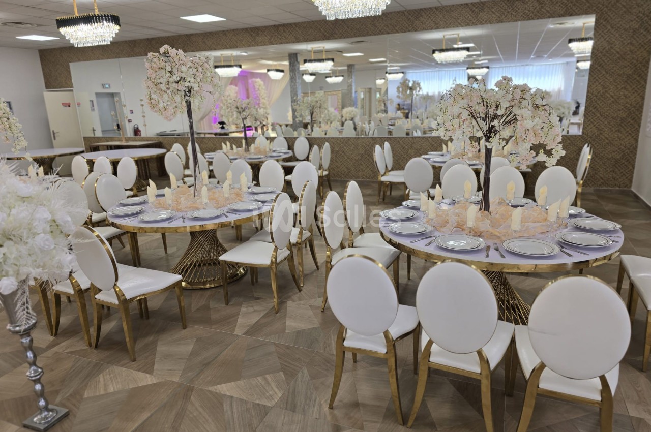 Salle de réception élégante avec tables rondes décorées, chaises blanches et lustres suspendus.