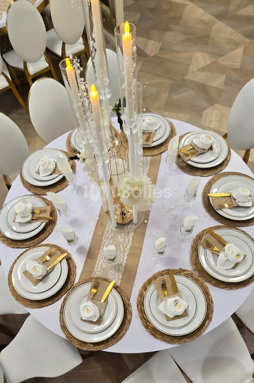 Table ronde élégamment dressée avec vaisselle blanche, serviettes dorées, bougies et décorations florales blanches.