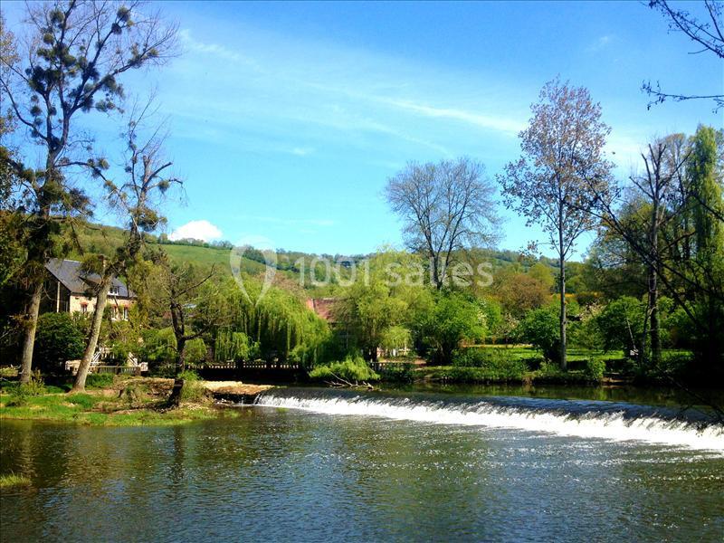 Paysage bucolique avec une rivière, petite cascade, arbres verdoyants et une maison en pierre sur la rive.
