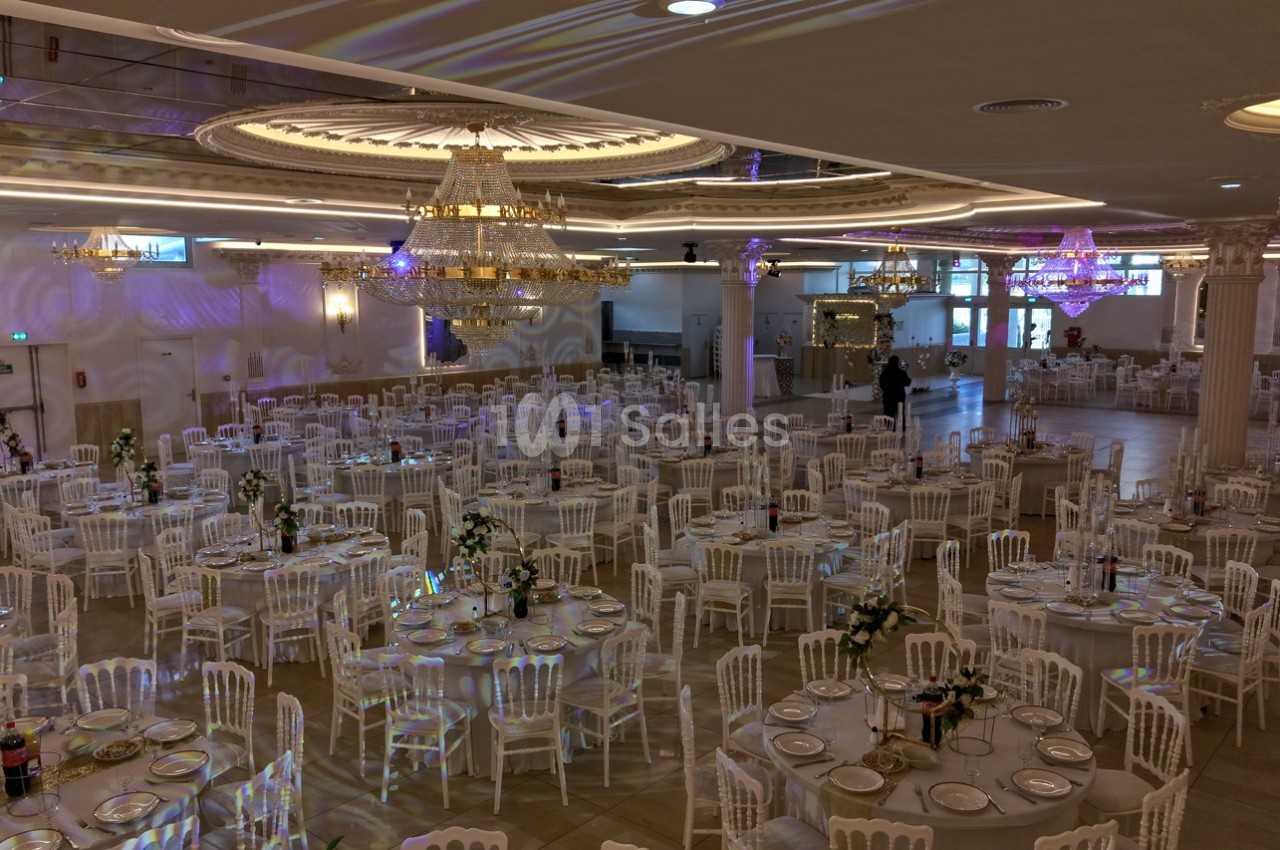Salle de réception élégante avec tables rondes dressées, chaises blanches et lustres imposants au plafond.
