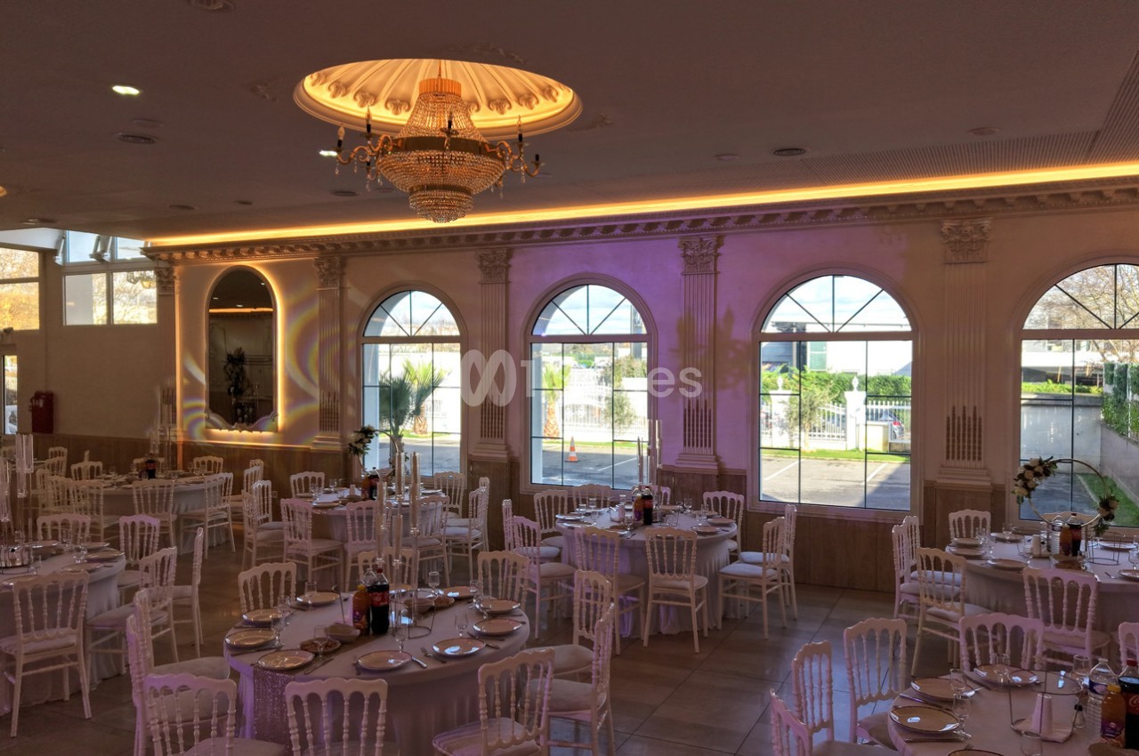 Salle de réception élégante avec tables dressées, chaises blanches et grandes fenêtres laissant entrer la lumière naturelle.