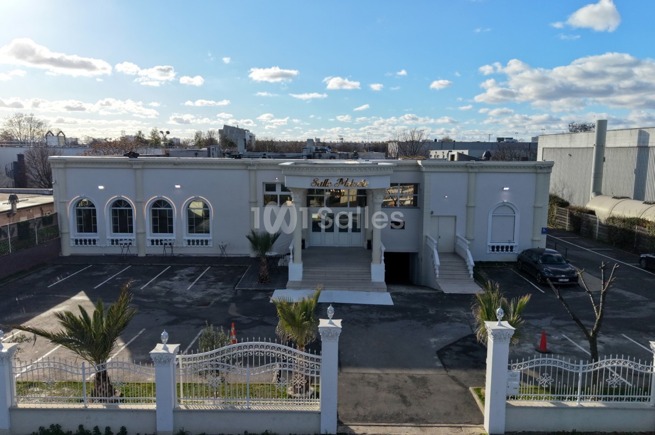 Façade d'un bâtiment avec une entrée principale, un parking et des palmiers, sous un ciel partiellement nuageux.