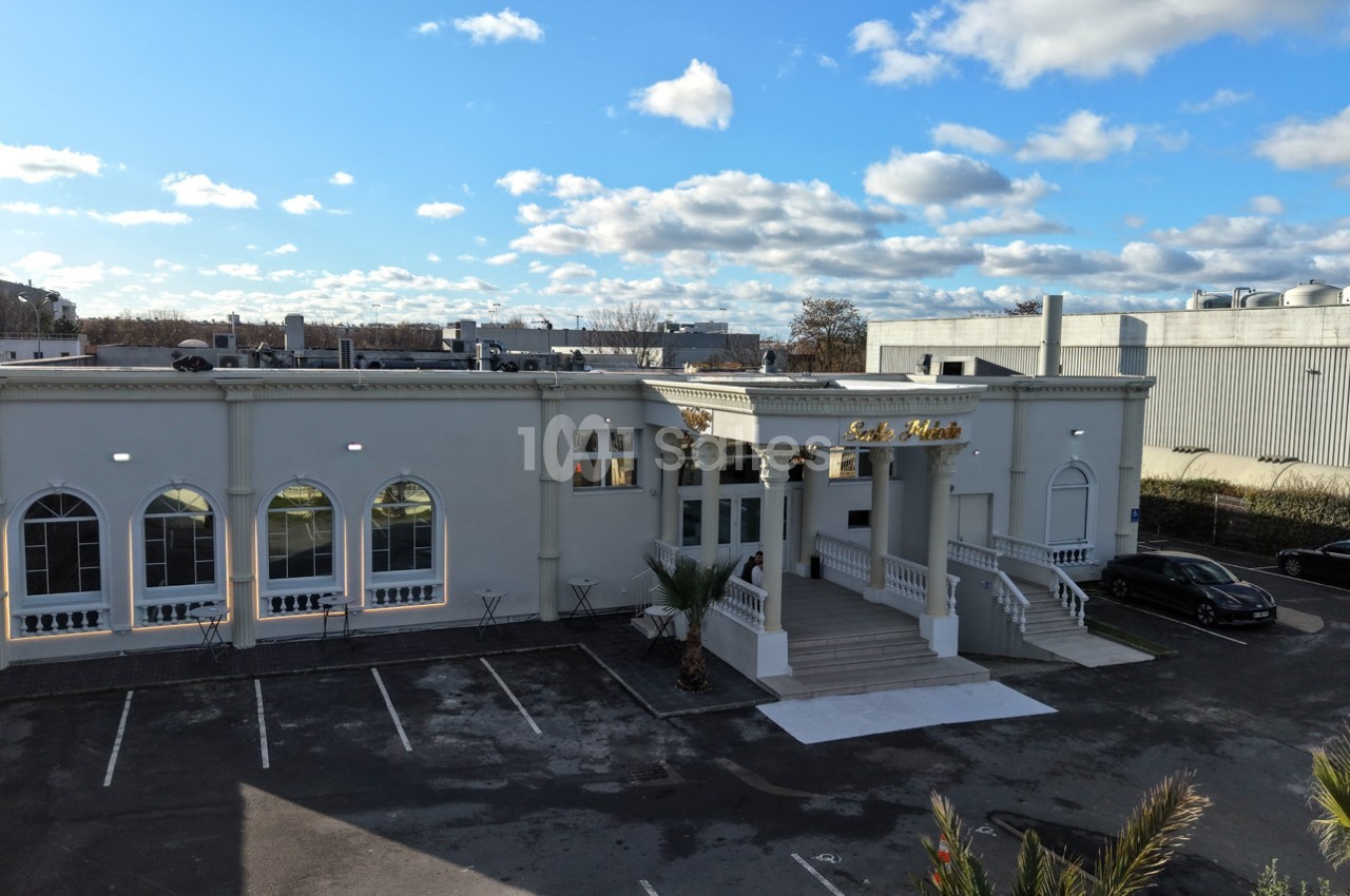 Façade d'un bâtiment blanc avec des arches vitrées, un escalier central et un parking vide à l'avant.