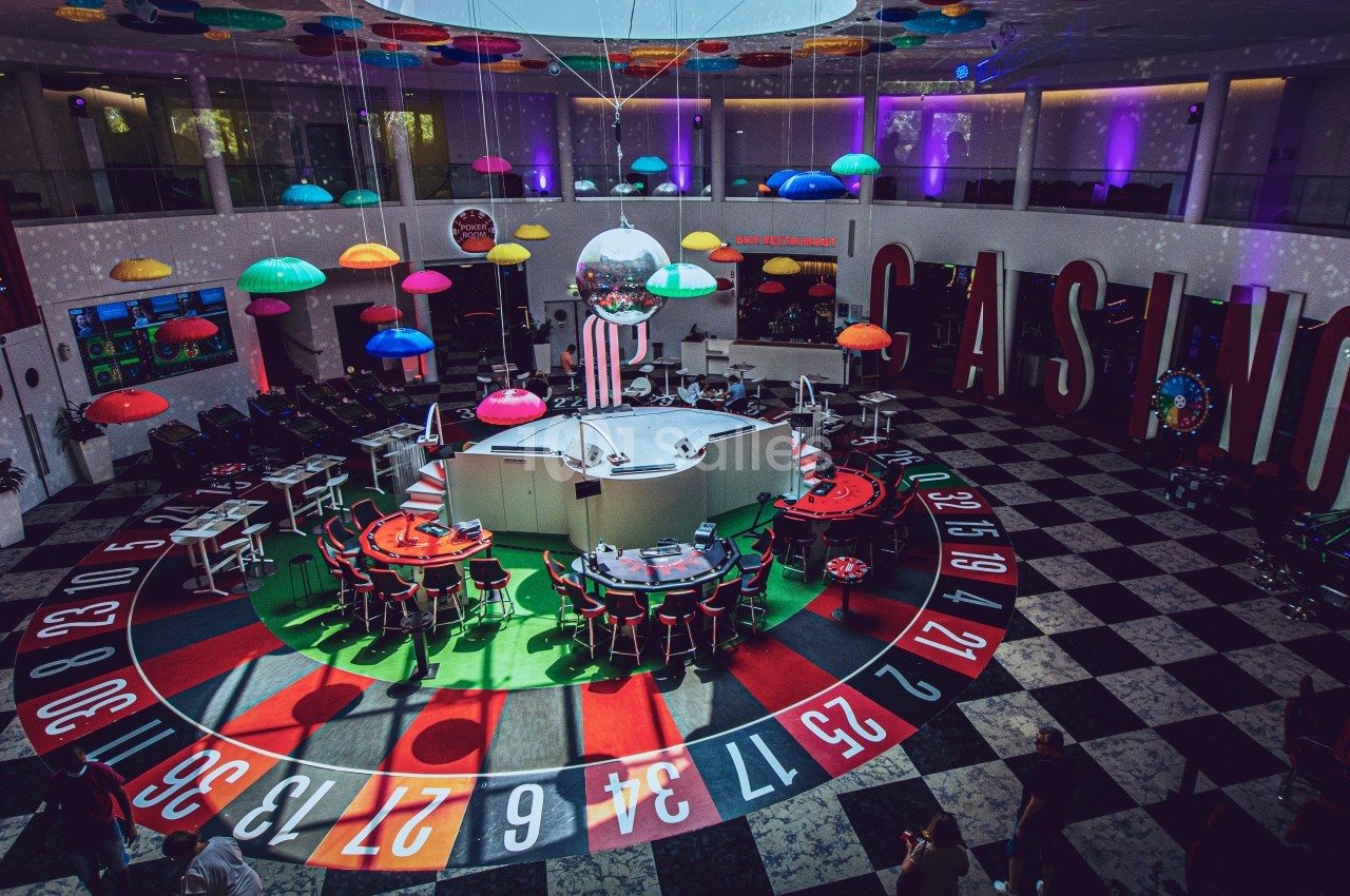Salle de casino colorée avec une grande roulette au sol, des tables de jeu et des lampes suspendues en forme de parapluies.