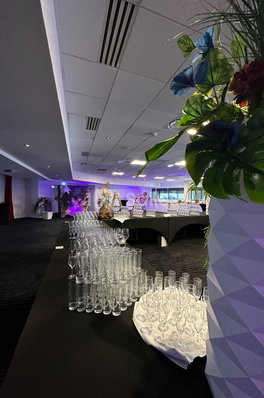 Salle de réception avec tables dressées, verres alignés au premier plan et décorations florales en arrière-plan.