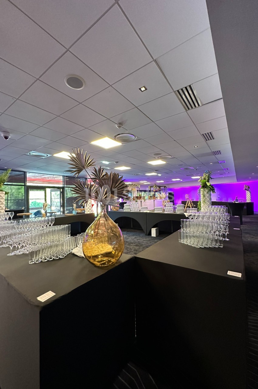 Salle de réception avec tables noires, verres disposés, décoration florale et éclairage violet en arrière-plan.