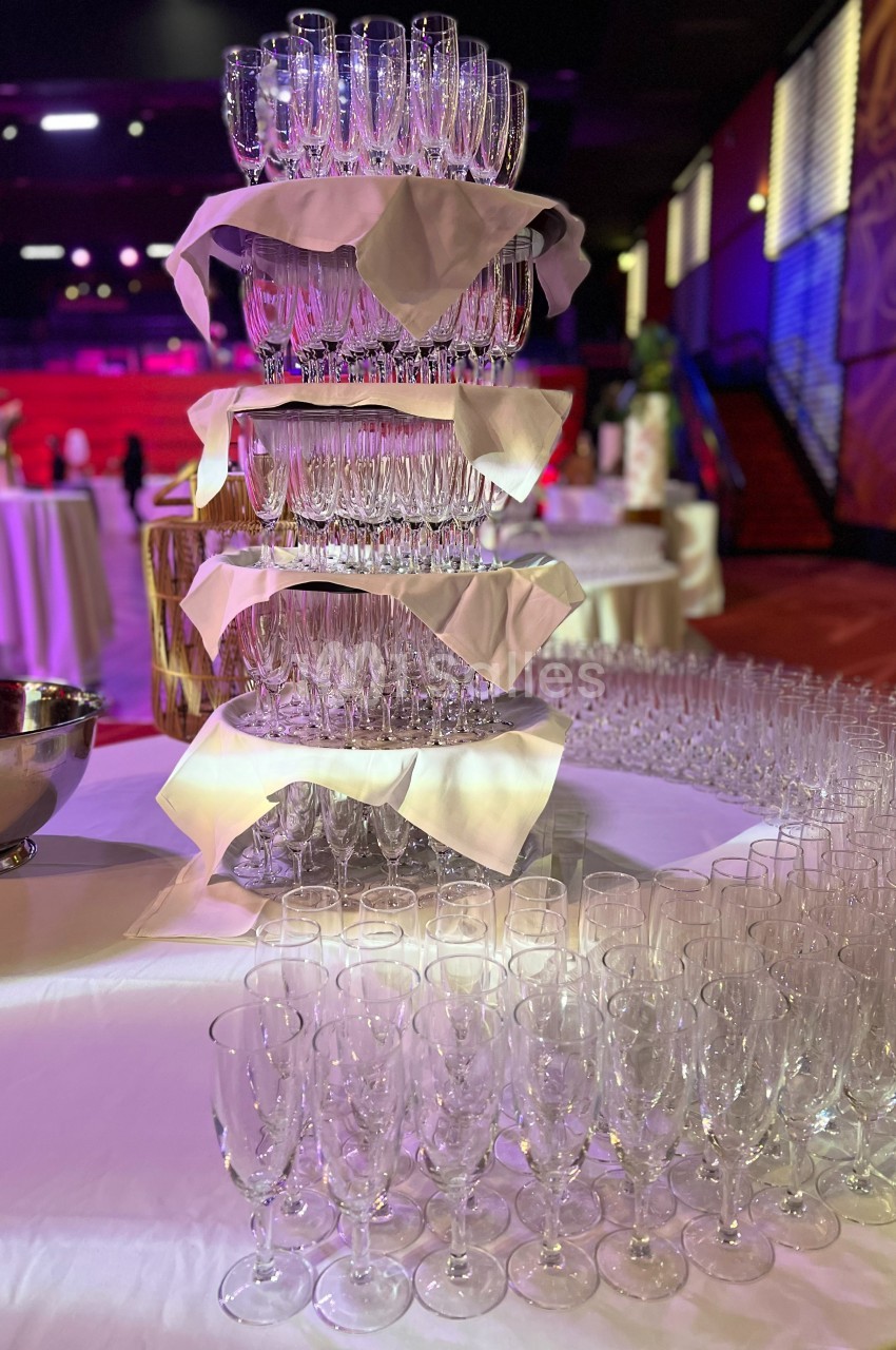 Tour de flûtes à champagne empilées avec des serviettes blanches, entourée de tables et de verres supplémentaires.