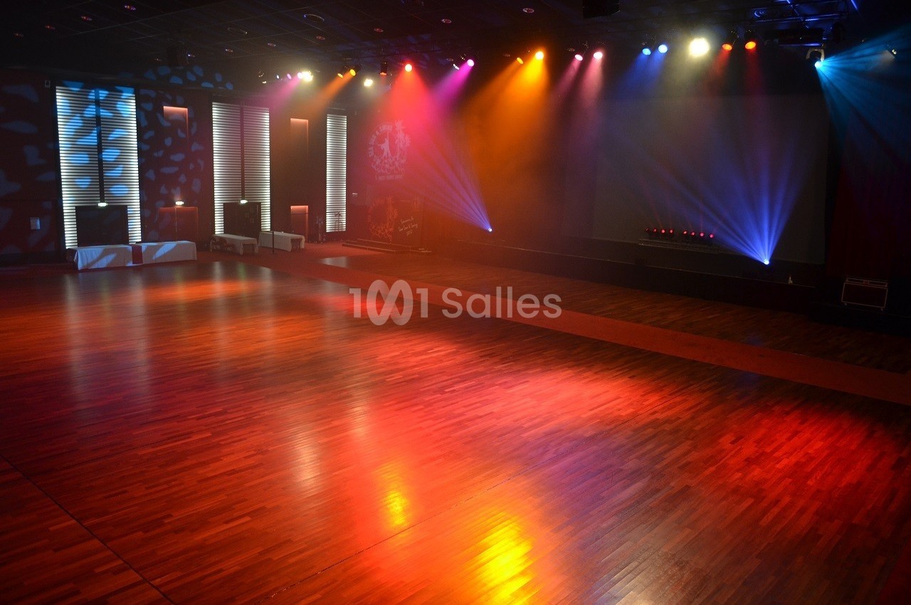 Salle de danse vide avec parquet en bois, éclairée par des projecteurs colorés et des lumières tamisées.