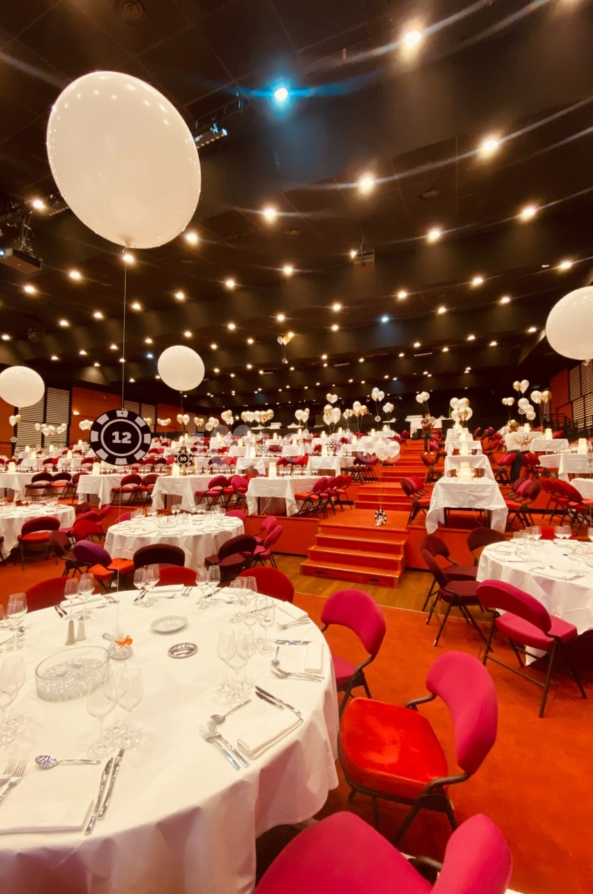 Salle de réception décorée avec des ballons blancs, des tables rondes dressées et des chaises rouges.