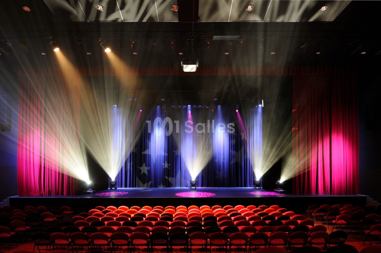 Salle de spectacle avec sièges rouges, scène éclairée par des projecteurs colorés et rideaux rouges ouverts.