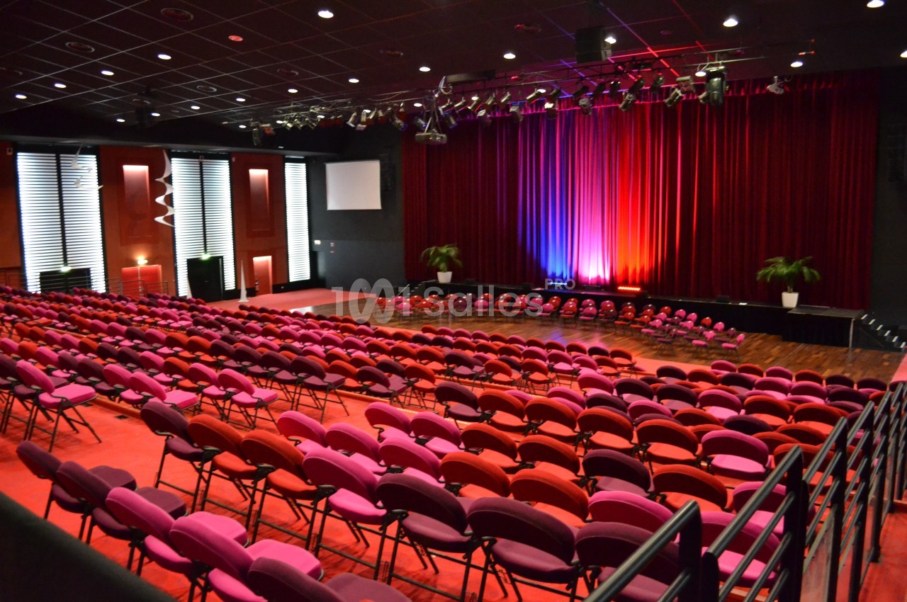 Salle de spectacle avec rangées de sièges rouges et roses, scène éclairée par des lumières colorées et rideaux rouges.