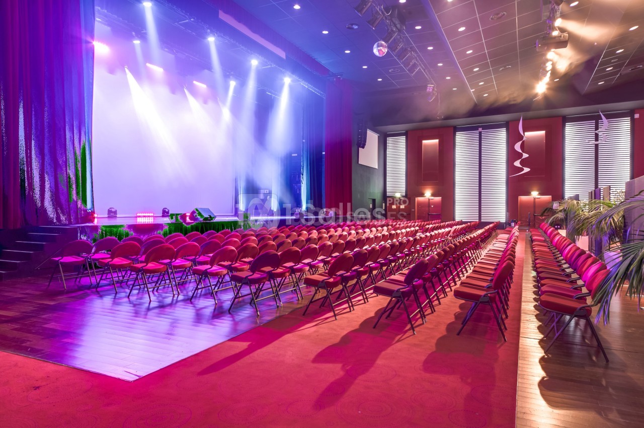 Salle de spectacle avec rangées de sièges rouges, scène éclairée par des projecteurs et rideaux ouverts.