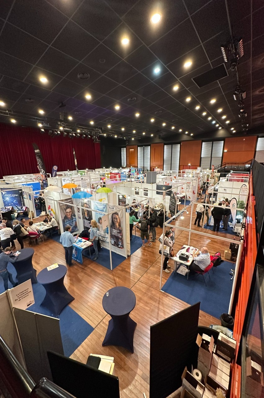 Vue d'ensemble d'un salon professionnel avec des stands, des visiteurs et une scène dans une grande salle éclairée.