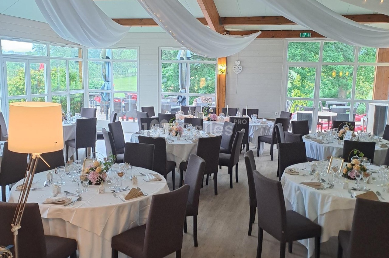 Salle de réception décorée avec des tables rondes dressées, nappes blanches, chaises noires et guirlandes suspendues.