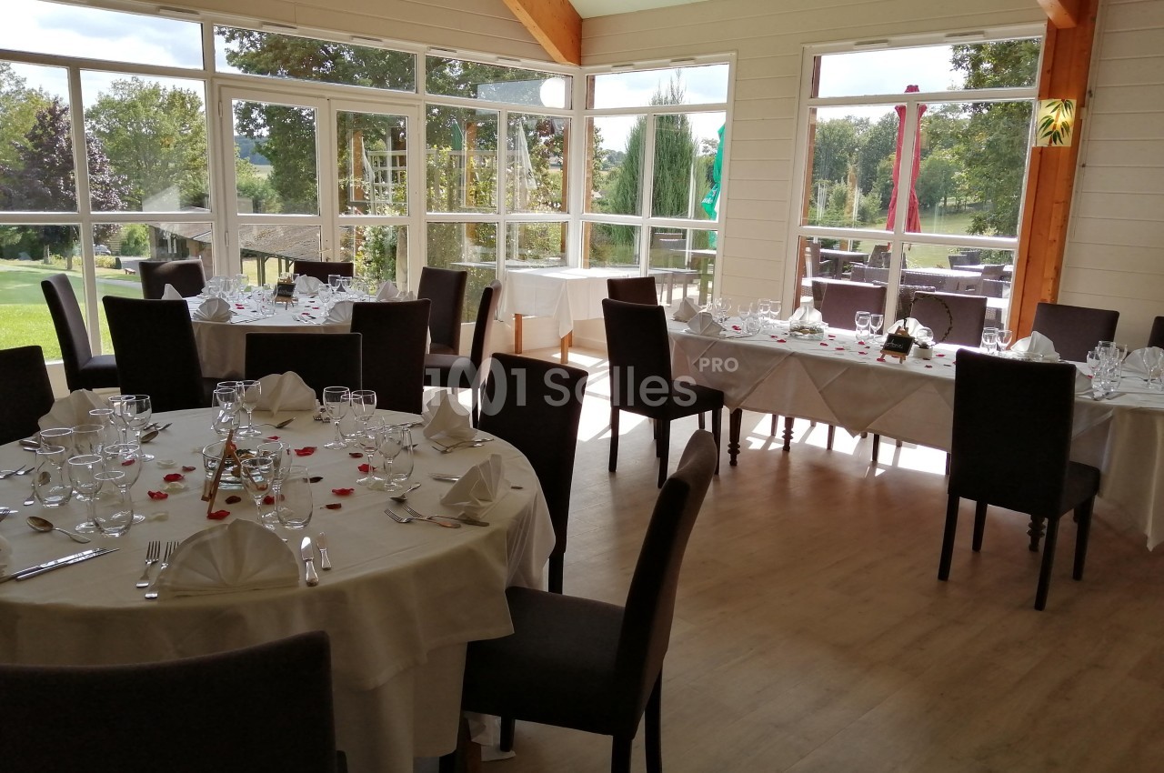 Salle lumineuse avec grandes baies vitrées, tables rondes et rectangulaires dressées pour un repas, vue sur un jardin.