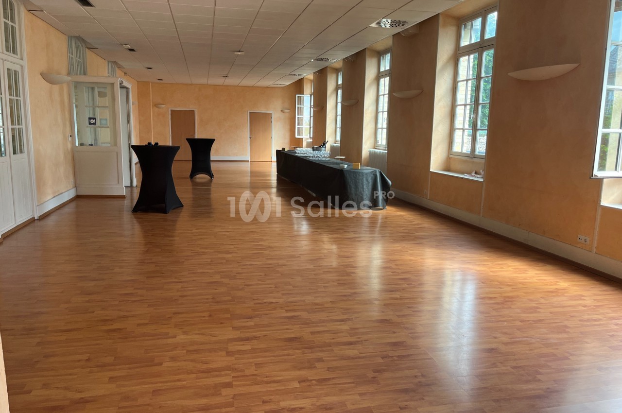 Salle lumineuse avec parquet, grandes fenêtres, murs clairs et tables hautes noires disposées le long d'un buffet.