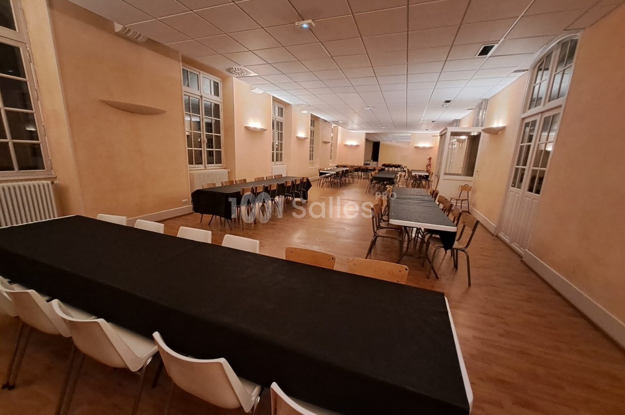 Salle spacieuse avec tables rectangulaires couvertes de nappes noires, chaises blanches et parquet clair.