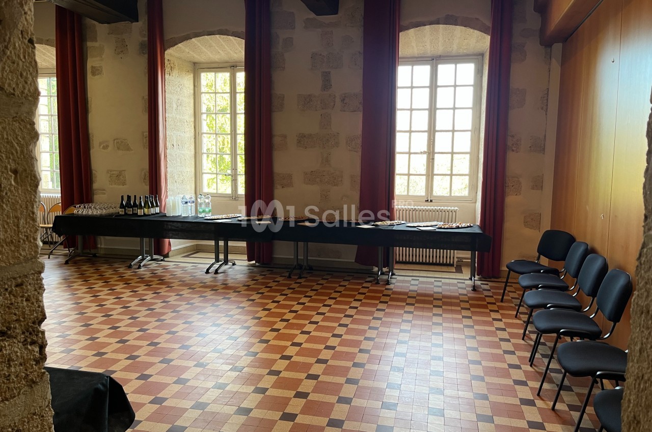 Salle lumineuse avec murs en pierre, sol en carreaux colorés, tables dressées avec boissons et chaises alignées.