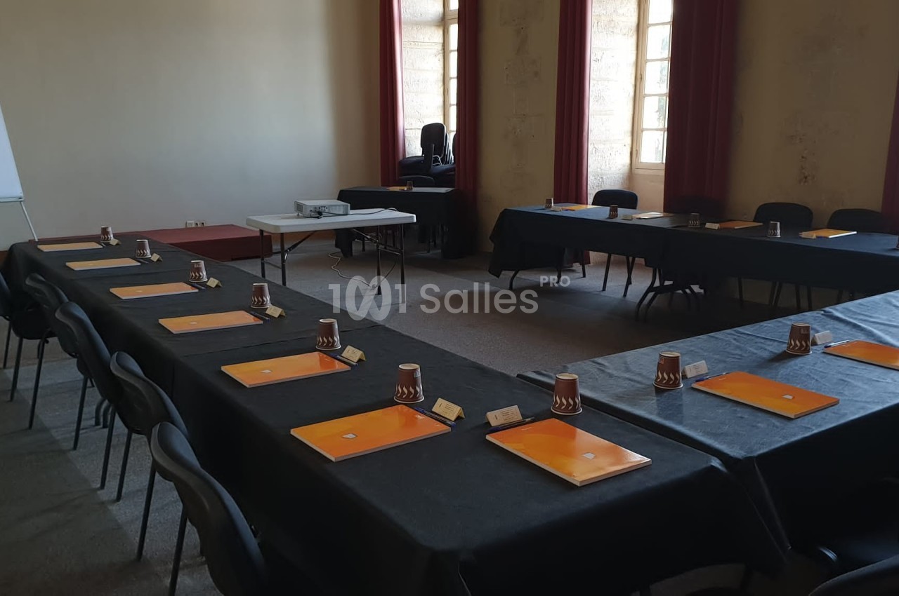 Salle de réunion avec tables disposées en U, équipées de dossiers, gobelets et un projecteur au fond de la pièce.