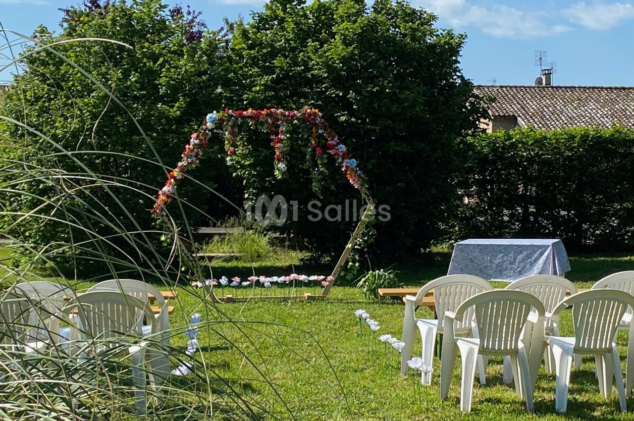 Chaises blanches disposées en extérieur devant une arche fleurie et une table recouverte d'une nappe, dans un jardin…