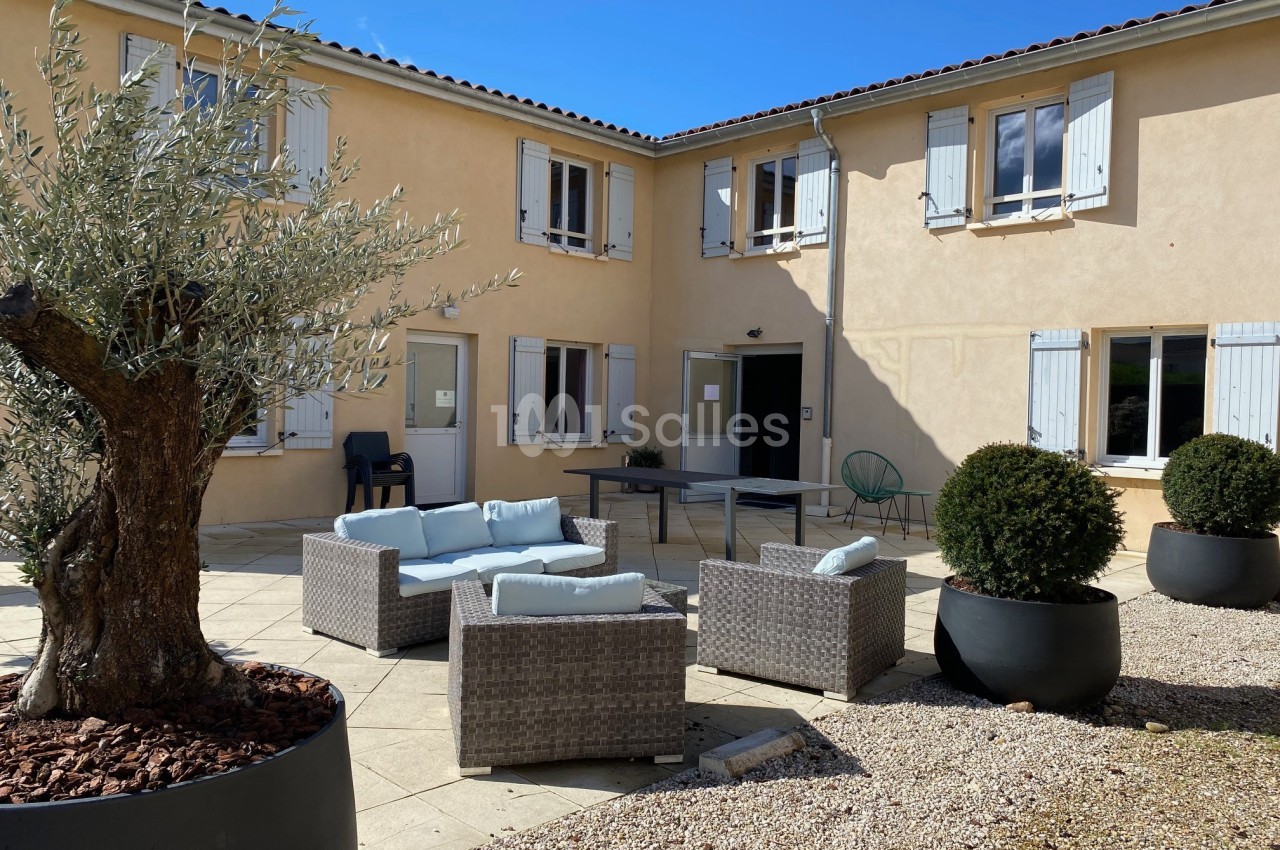 Cour intérieure avec mobilier d'extérieur en rotin, olivier et arbustes en pots, entourée d'un bâtiment beige à volets…