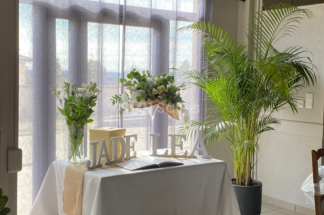 Table décorée avec des fleurs, des plantes en pot et les lettres ’JADE LEA’ devant une fenêtre avec rideaux translucides.
