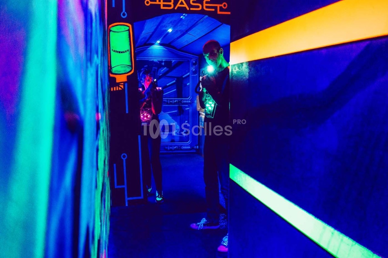 Deux personnes jouent dans une salle de laser game éclairée par des lumières fluorescentes.
