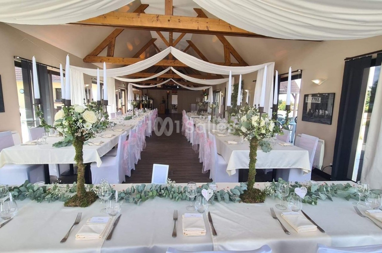 Salle de réception décorée avec des tables alignées, nappes blanches, chandeliers et compositions florales élégantes.