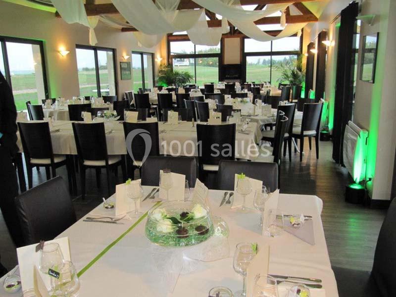 Salle de réception décorée avec des tables dressées, nappes blanches et éclairage naturel provenant de grandes baies vitrées.