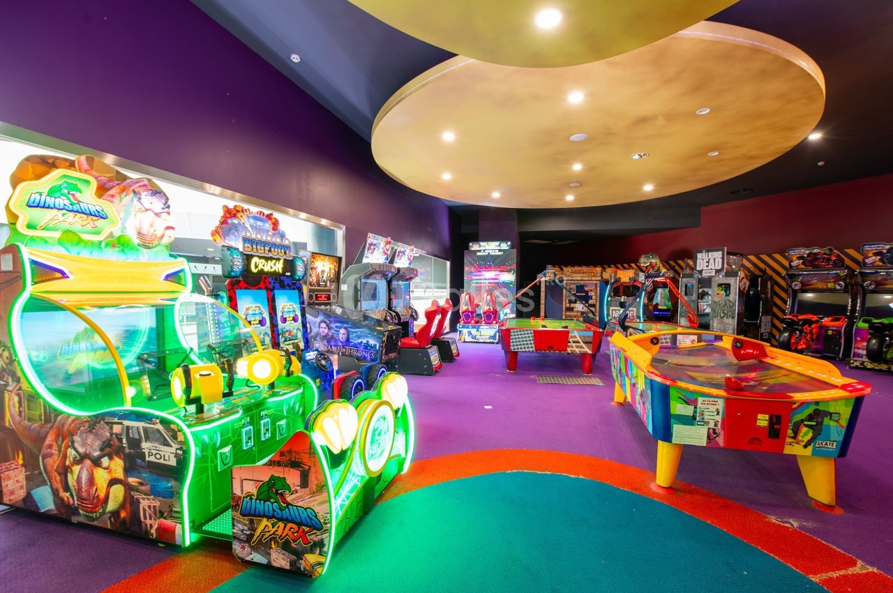 Salle d'arcade colorée avec divers jeux vidéo, machines interactives et une table de air hockey sur un sol violet.