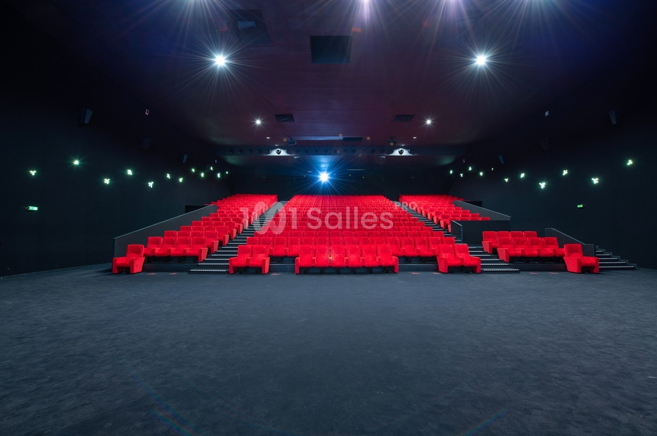 Salle de cinéma vide avec des rangées de fauteuils rouges et un écran illuminé en arrière-plan.