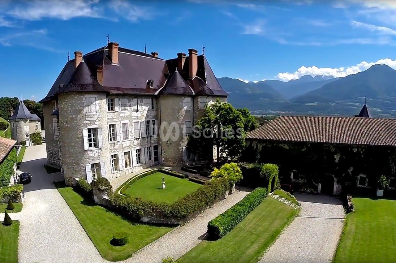 Château en pierre entouré de jardins verdoyants, avec vue sur des montagnes sous un ciel bleu dégagé.