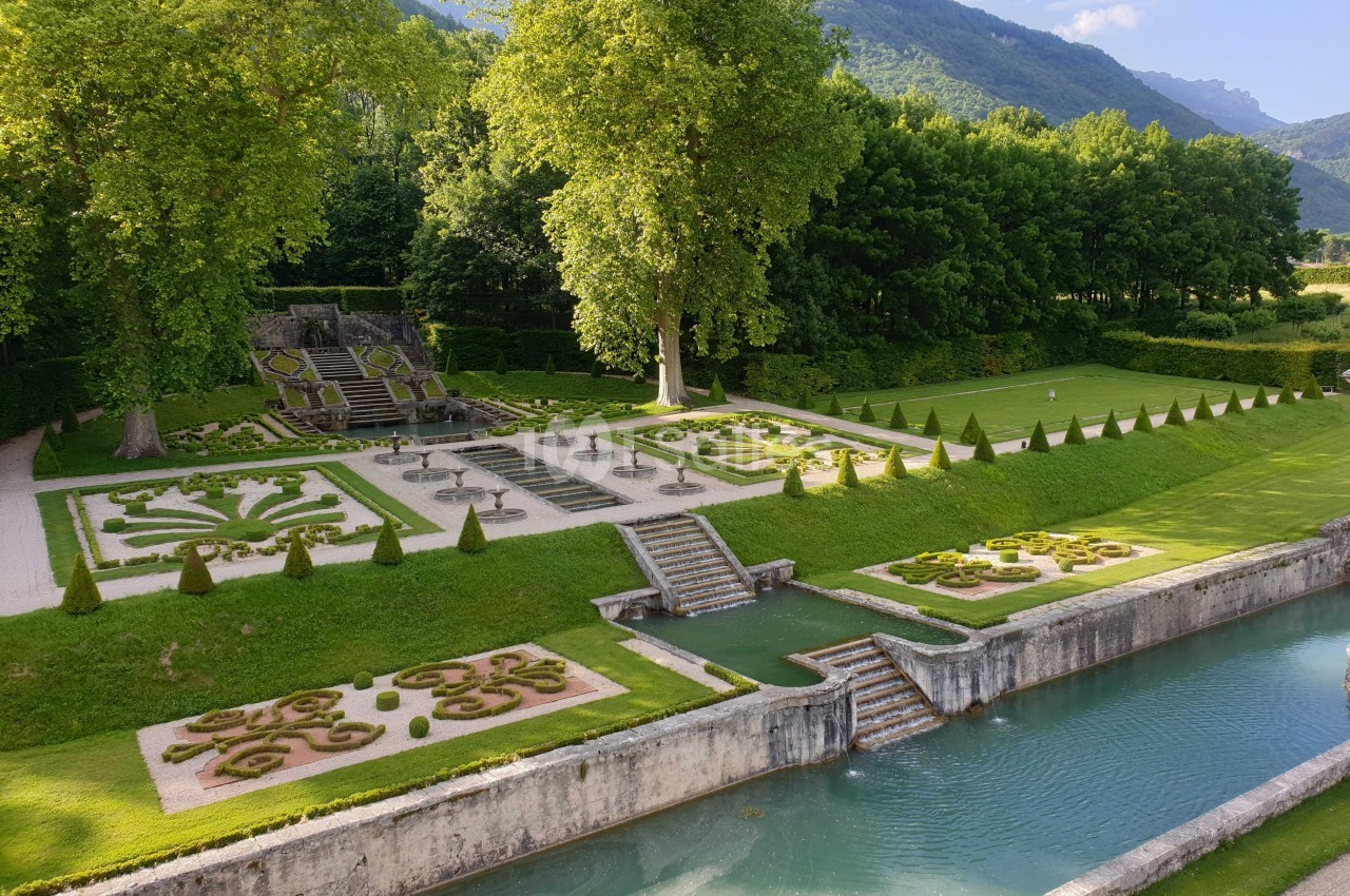 Jardin à la française avec parterres symétriques, escaliers, canal et arbres, entouré de collines verdoyantes.