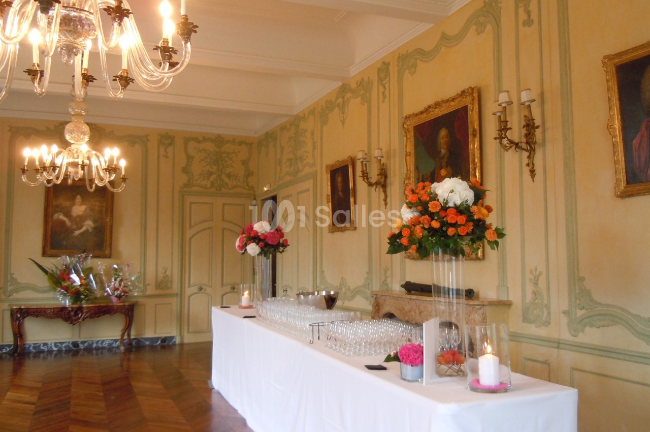 Salle élégante avec moulures, lustres, peintures anciennes et une table dressée avec verres et décorations florales.