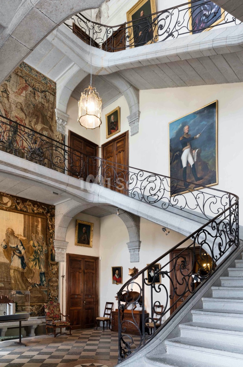 Escalier intérieur en pierre avec balustrades en fer forgé, entouré de tableaux, tapisseries et portes en bois.