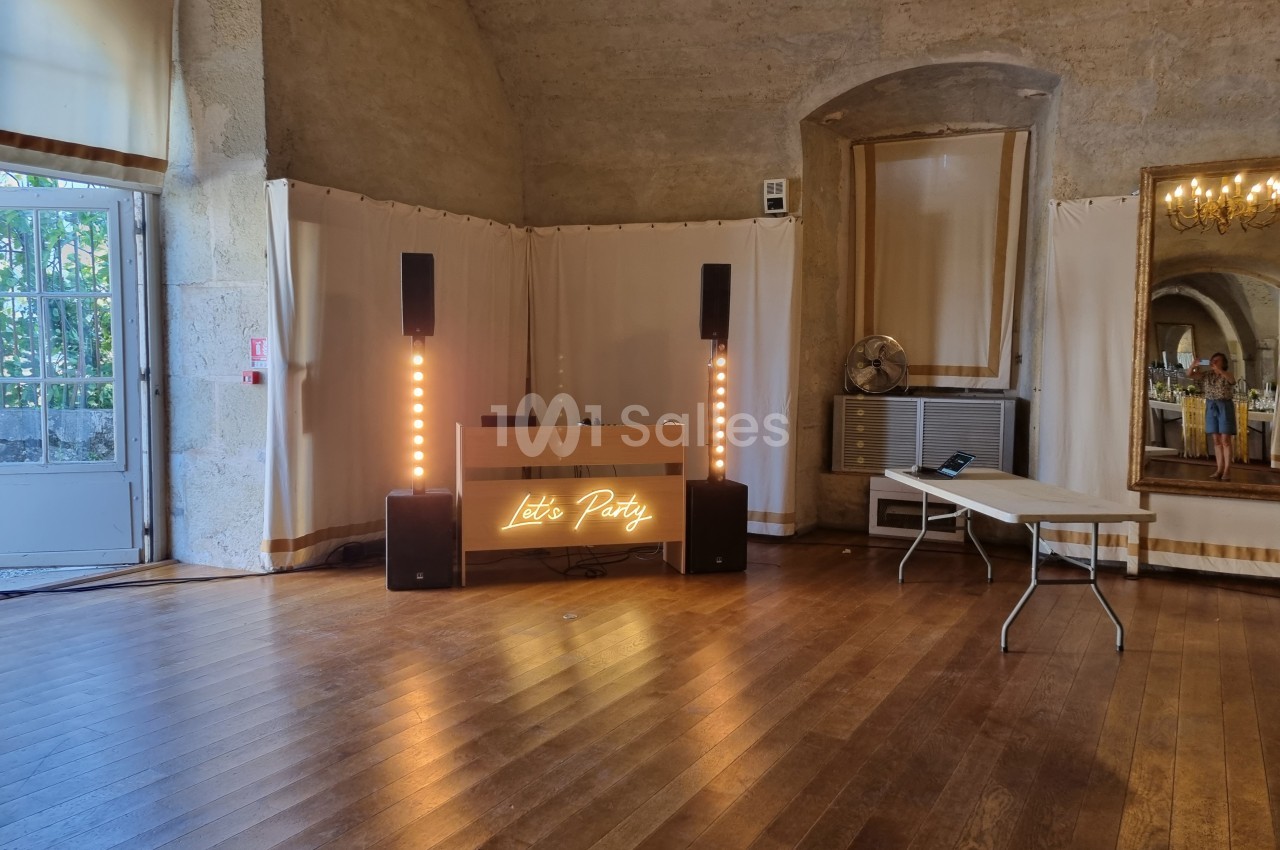 Salle avec parquet, table de mixage éclairée ’Let's Party’, enceintes et rideaux blancs, dans un cadre intérieur en pierre.