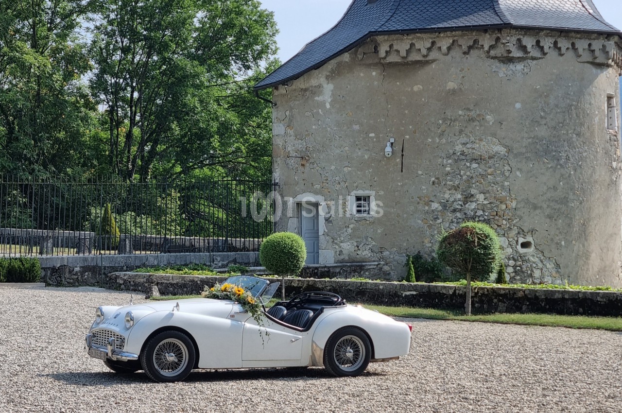 Voiture ancienne blanche décorée de fleurs, stationnée devant un bâtiment en pierre avec un toit en ardoise.