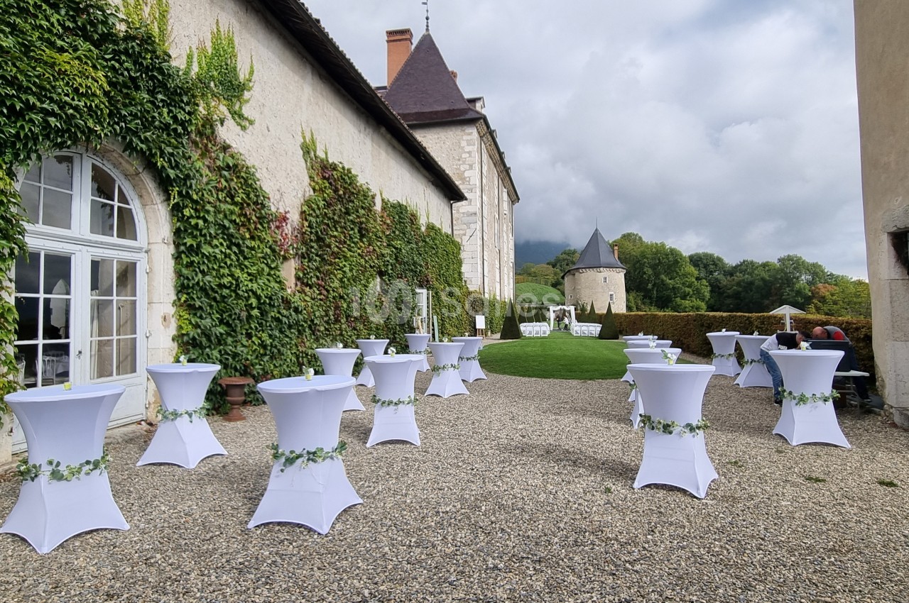 Cour extérieure d'un bâtiment ancien avec des tables hautes décorées de housses blanches et de guirlandes végétales.