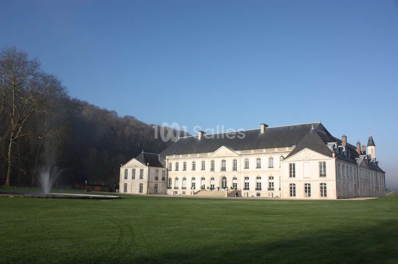 Façade d'un grand bâtiment historique en pierre, entouré d'une pelouse et d'une fontaine, sous un ciel dégagé.