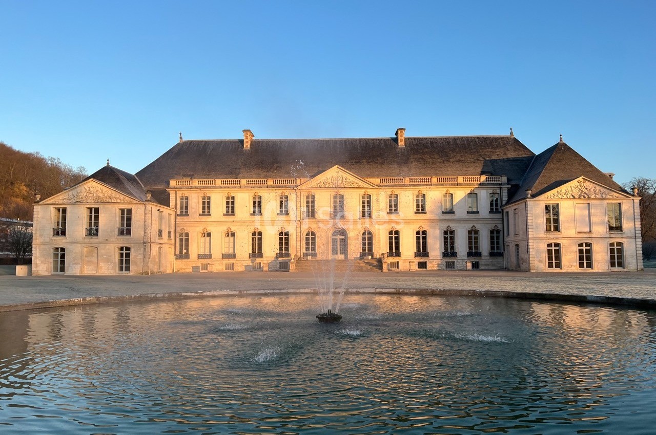Façade d'un château éclairé par le soleil couchant, avec une fontaine au centre d'un bassin au premier plan.