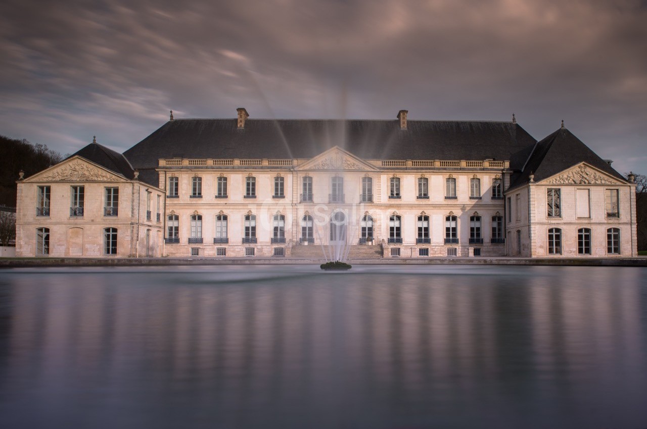 Façade d'un château élégant avec un bassin et une fontaine au premier plan, sous un ciel nuageux.