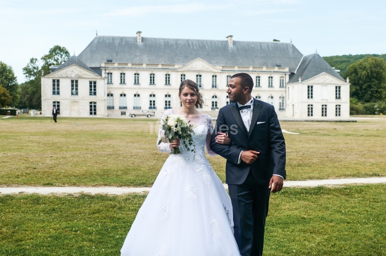 Un couple en tenue de mariage marche dans un parc devant un grand bâtiment historique.