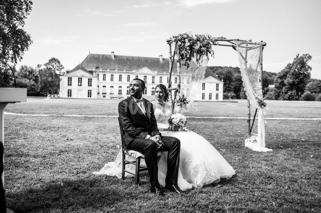 Un couple assis sous une arche décorée, dans un jardin devant un grand bâtiment historique.
