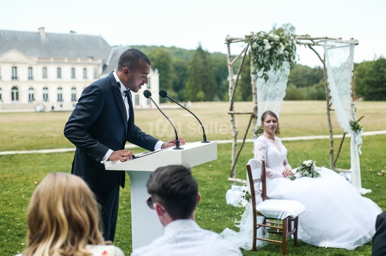 Un homme en costume prononce un discours devant un pupitre lors d'une cérémonie de mariage en extérieur.