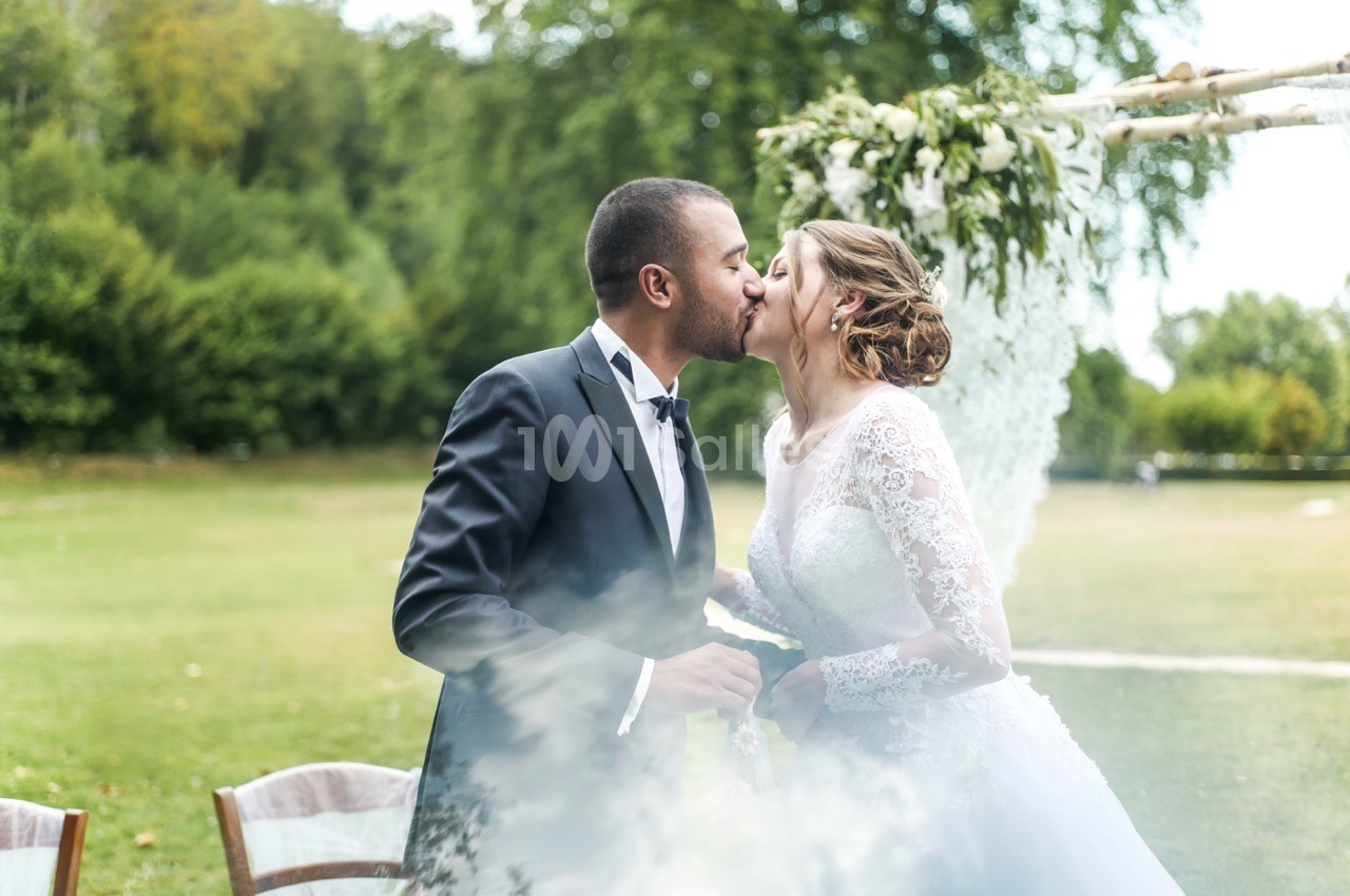 Un couple en tenue de mariage s'embrasse dans un jardin, sous une arche décorée de fleurs blanches.