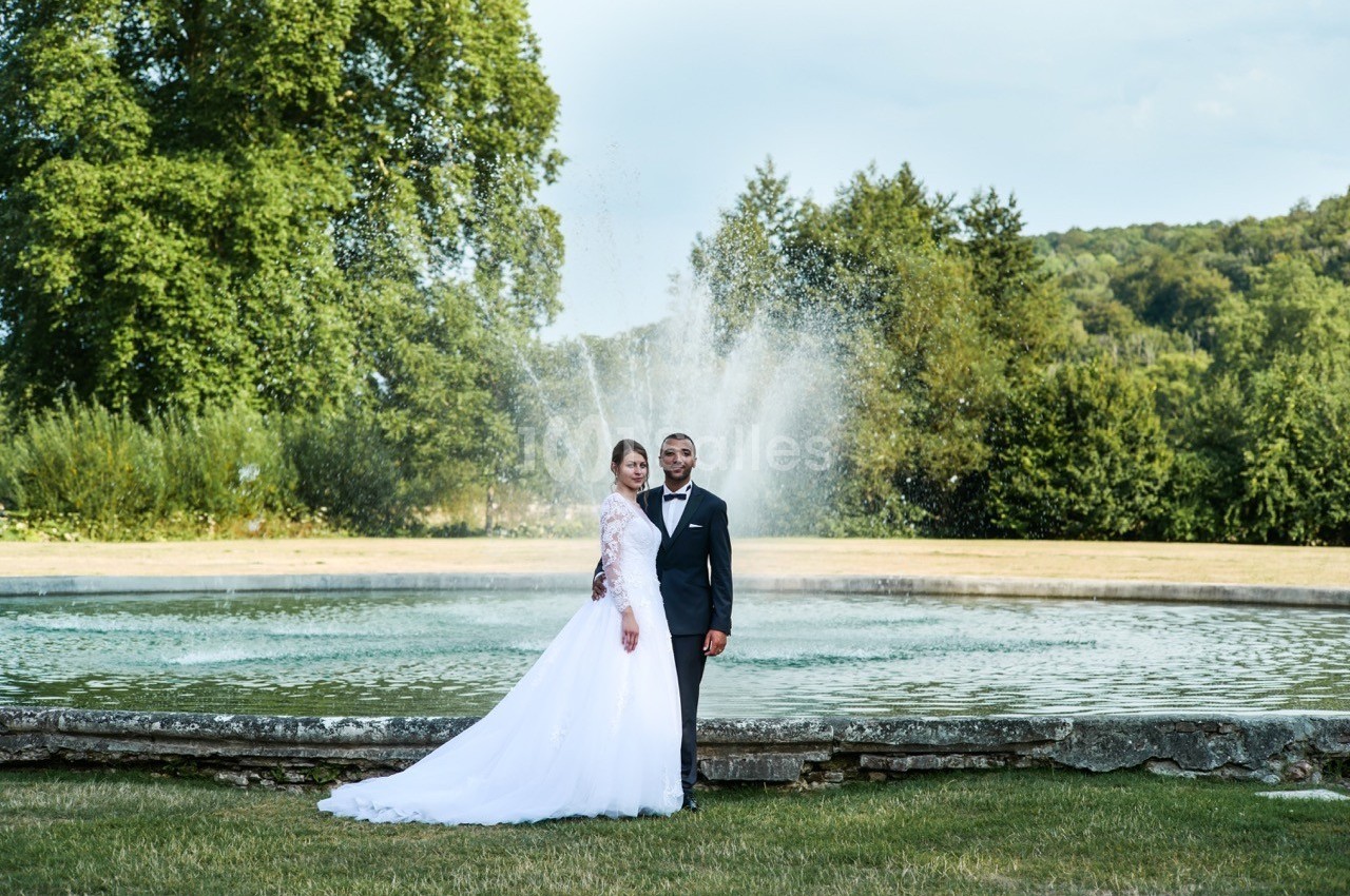 Un couple en tenue de mariage pose devant une fontaine dans un parc verdoyant.