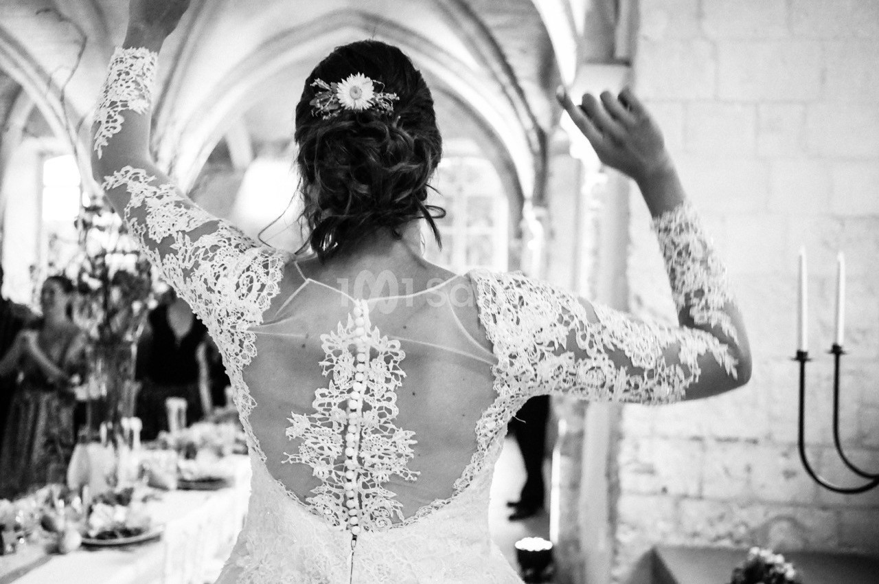 Une mariée de dos levant les bras, portant une robe en dentelle dans une salle voûtée décorée pour une réception.