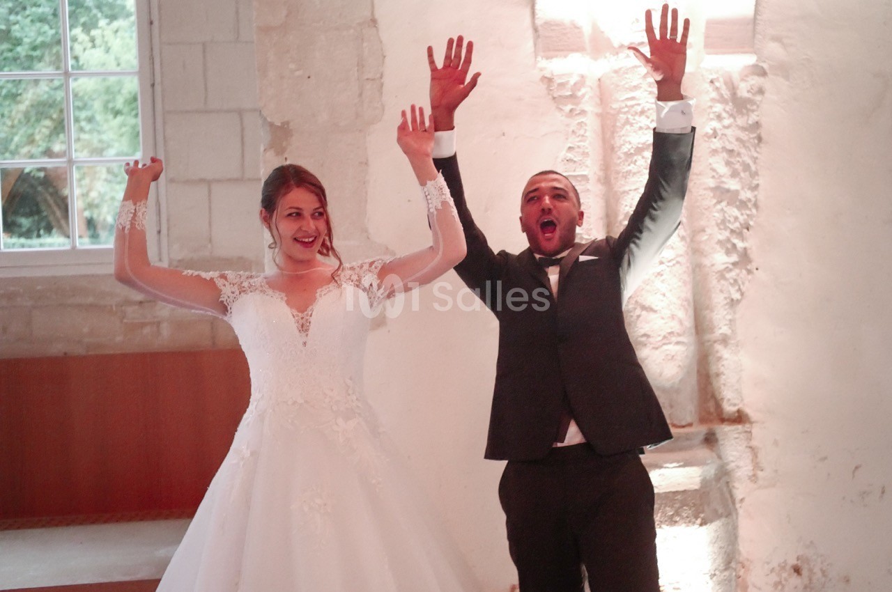 Un couple en tenue de mariage lève les bras en souriant dans une salle lumineuse aux murs en pierre.