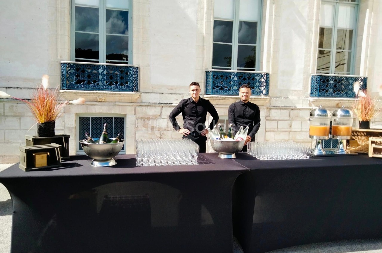 Deux serveurs en tenue noire derrière une table dressée avec boissons, verres et décorations, devant un bâtiment ancien.